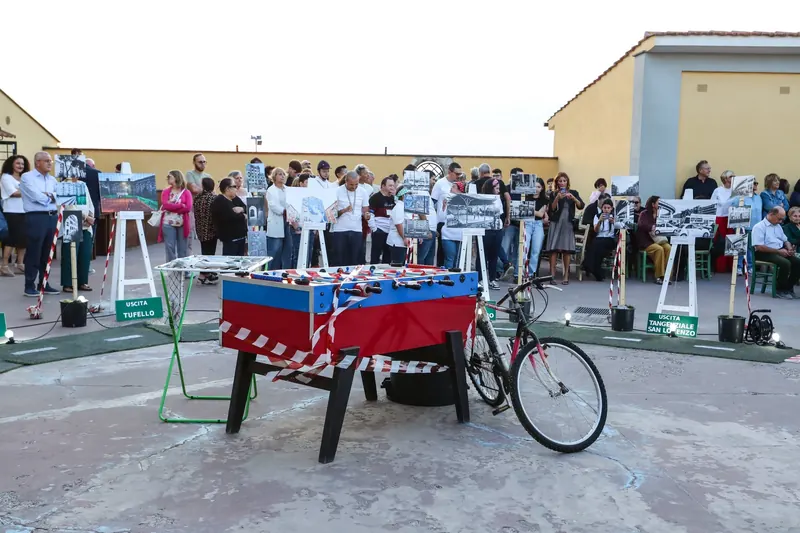 Un calcio balilla e una bicicletta avvolti nel nastro bianco e rosso, al centro del percorso, con il pubblico sullo sfondo